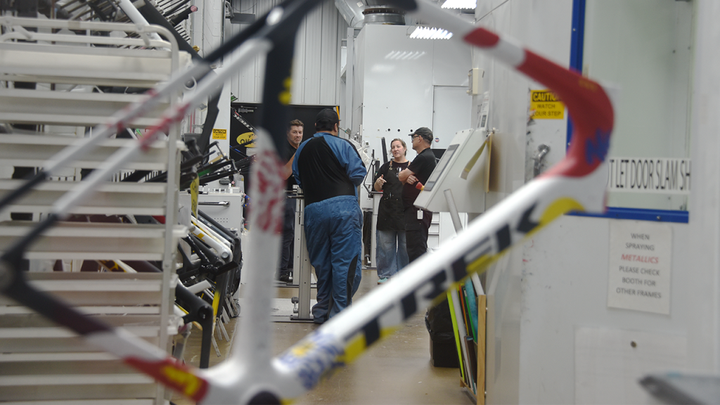 A Trek bike frame drying in the paint room while workers speak in a small group in the background