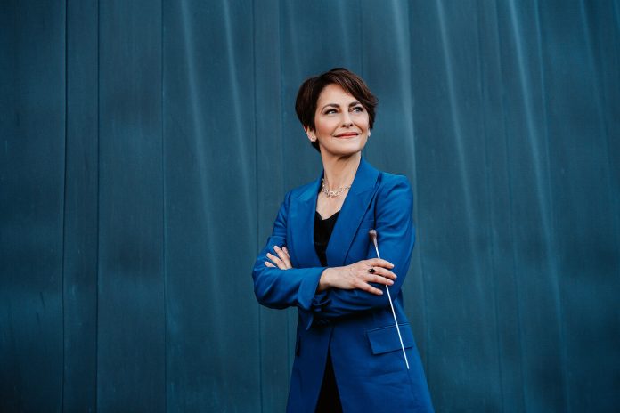 A woman donning a blue business coat stands with arms crossed and a conductor's wand