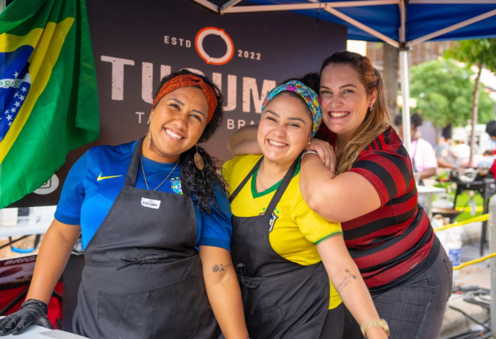 Three people in cooking attire pose at a food booth outside