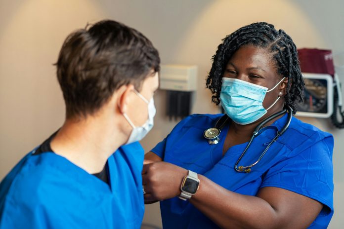 A nurse in blue scrubs checks the blood pressure of another nurse in blue scrubs