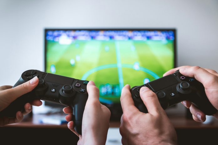 Two hands holding up PlayStation 4 controllers with the FIFA video game in the background