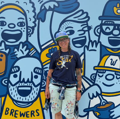 A woman standing in front of a blue and yellow Milwaukee Brewers mural she painted
